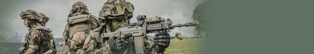 Drei Personen in Tarnkleidung in Stellung, die vorderste im Anschlag mit Sturmgewehr