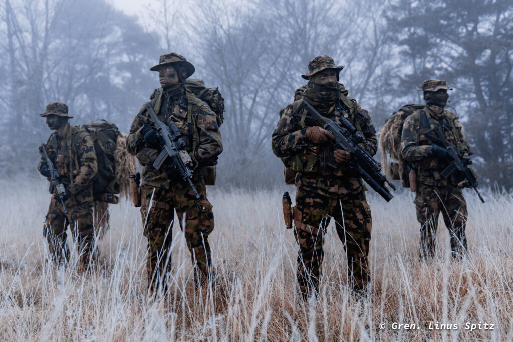 Vier Personen in Tarnkleidung mit Sturmgewehr bewegen sich bei Frost und Nebel durch hohes Gras